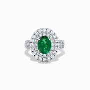 The Brasilica 18K White Gold Emerald and Diamond Double Halo Ring features an oval green emerald encircled by two rows of diamonds and delicate side stones, all set on a white gold band against a white background.