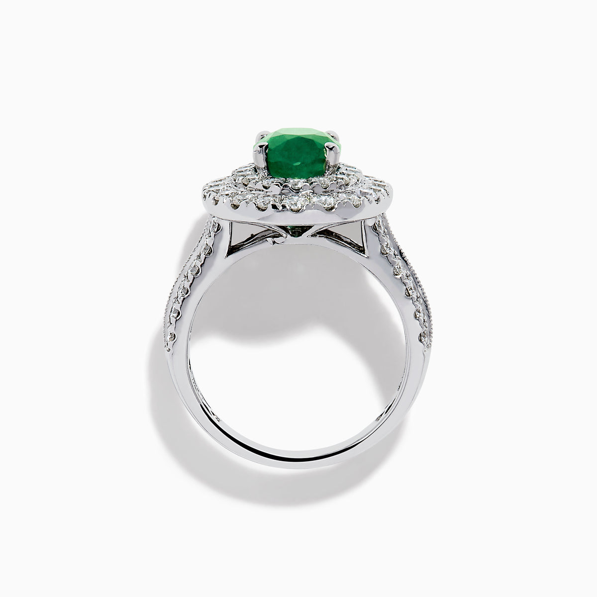 The Brasilica 18K White Gold Emerald and Diamond Double Halo Ring features an oval green emerald encircled by two rows of diamonds and delicate side stones, all set on a white gold band against a white background.