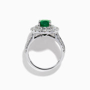 The Brasilica 18K White Gold Emerald and Diamond Double Halo Ring features an oval green emerald encircled by two rows of diamonds and delicate side stones, all set on a white gold band against a white background.