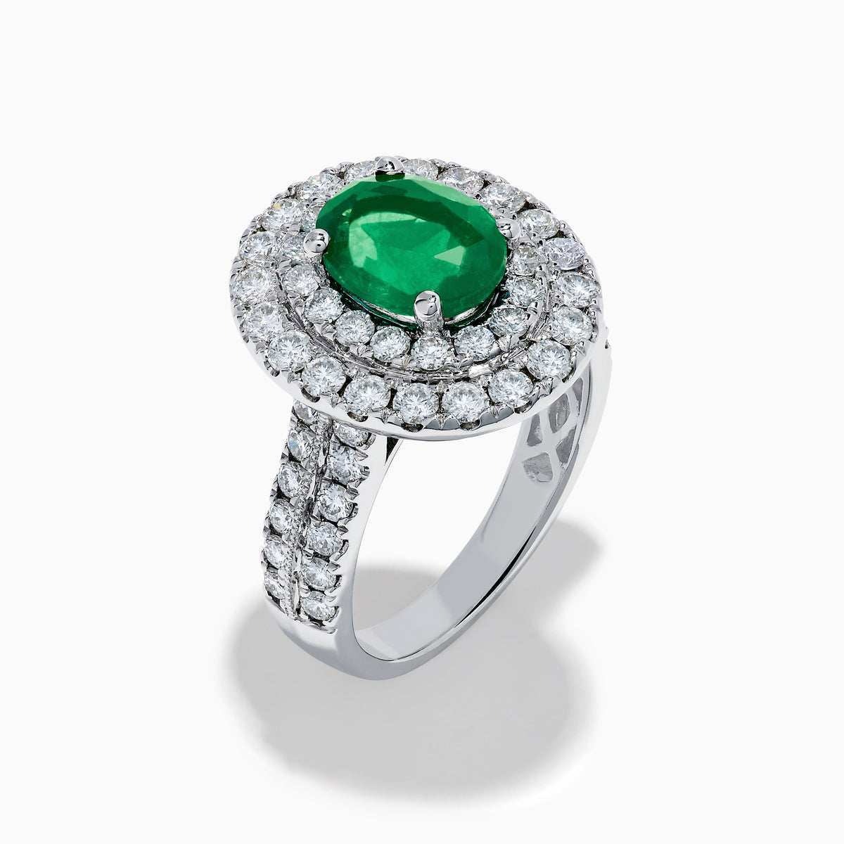 The Brasilica 18K White Gold Emerald and Diamond Double Halo Ring features an oval green emerald encircled by two rows of diamonds and delicate side stones, all set on a white gold band against a white background.