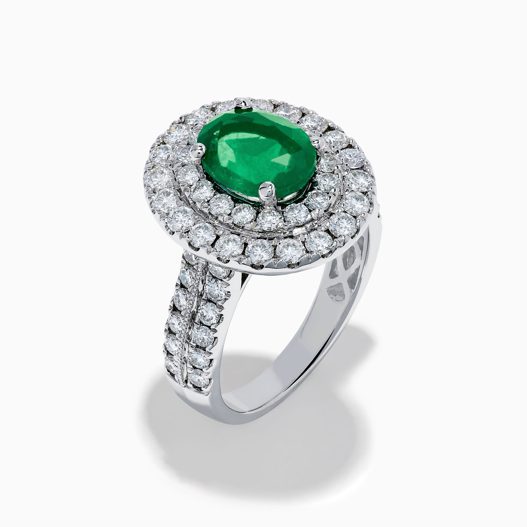 The Brasilica 18K White Gold Emerald and Diamond Double Halo Ring features an oval green emerald encircled by two rows of diamonds and delicate side stones, all set on a white gold band against a white background.
