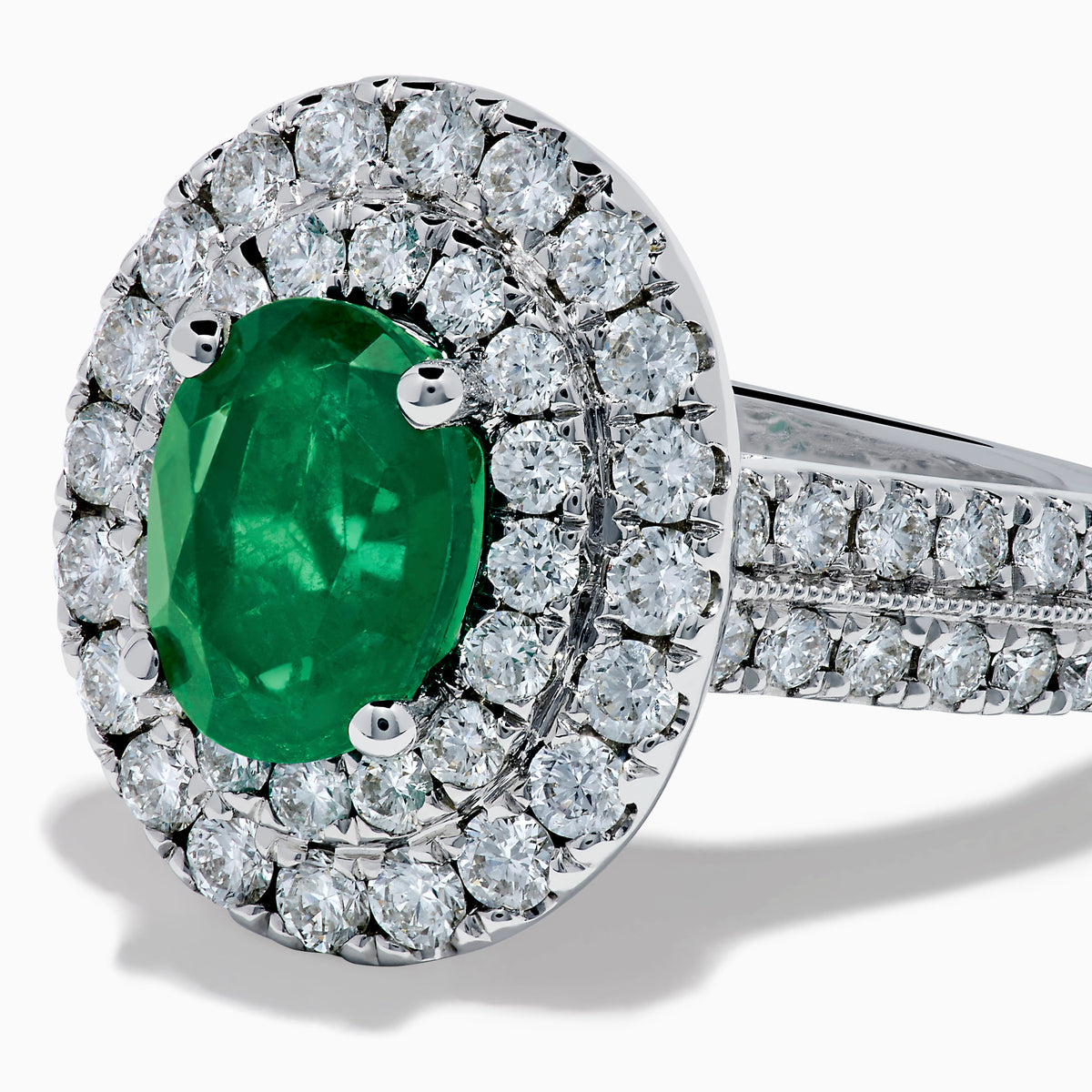 The Brasilica 18K White Gold Emerald and Diamond Double Halo Ring features an oval green emerald encircled by two rows of diamonds and delicate side stones, all set on a white gold band against a white background.