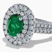 The Brasilica 18K White Gold Emerald and Diamond Double Halo Ring features an oval green emerald encircled by two rows of diamonds and delicate side stones, all set on a white gold band against a white background.