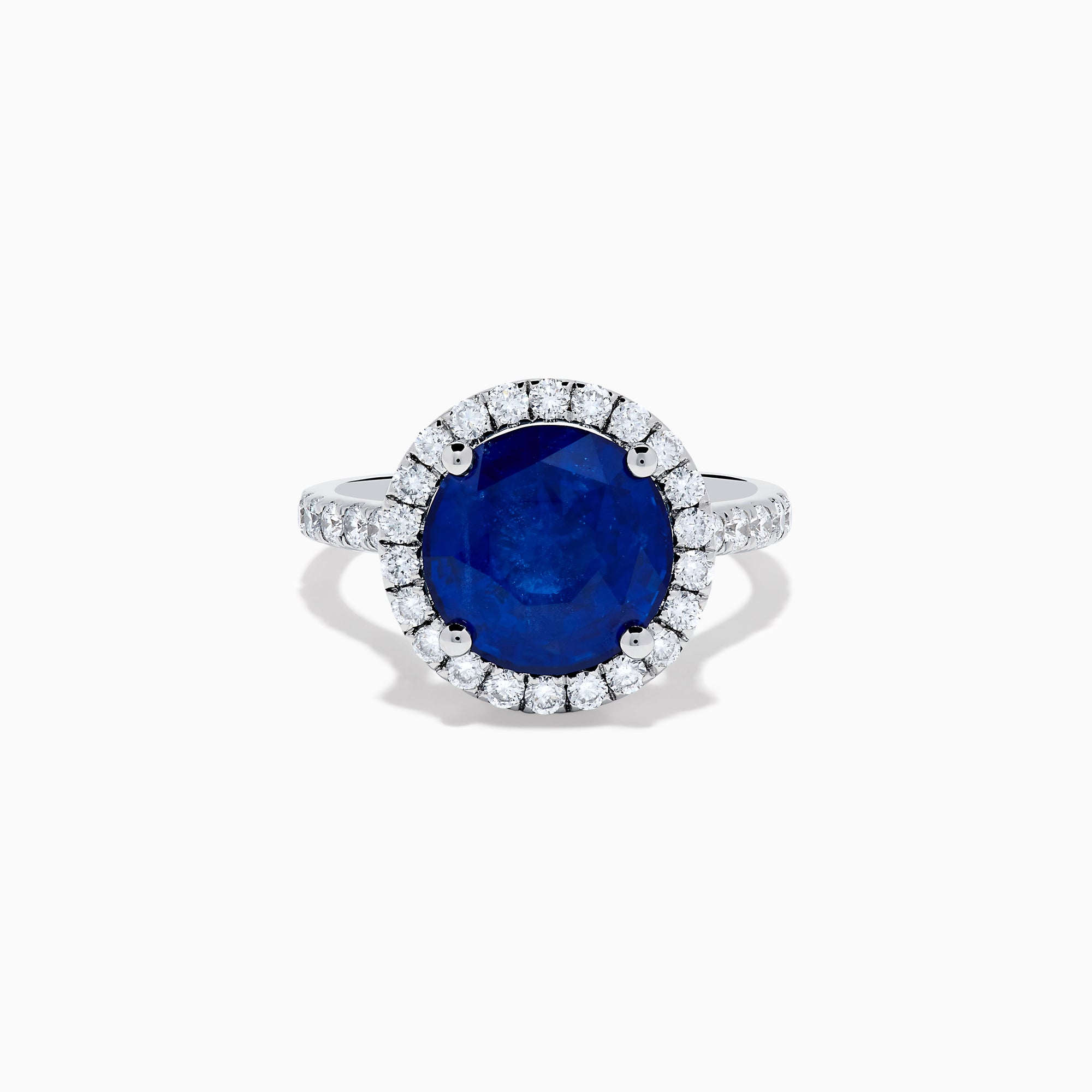 The Royale Blue 18K White Gold Blue Sapphire and Diamond Ring features a striking round blue sapphire center, encircled by a halo of sparkling diamonds, displayed against a white background.