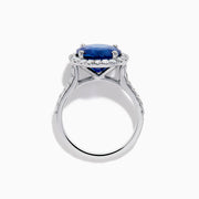The Royale Blue 18K White Gold Blue Sapphire and Diamond Ring features a striking round blue sapphire center, encircled by a halo of sparkling diamonds, displayed against a white background.
