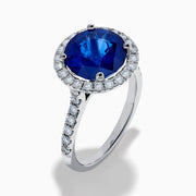 The Royale Blue 18K White Gold Blue Sapphire and Diamond Ring features a striking round blue sapphire center, encircled by a halo of sparkling diamonds, displayed against a white background.