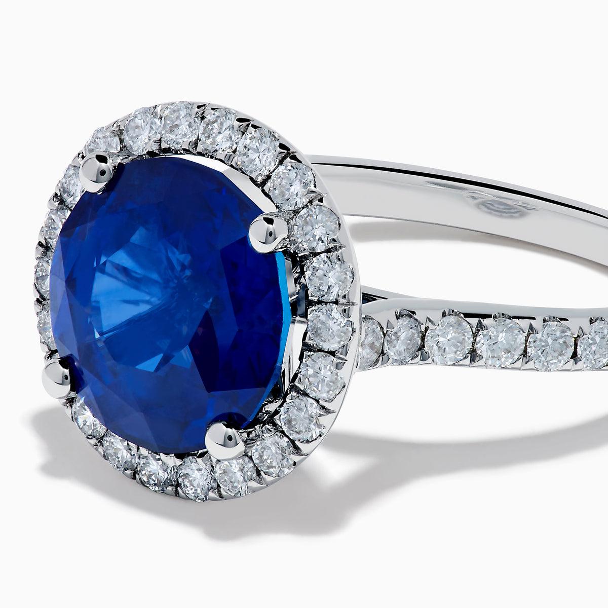 The Royale Blue 18K White Gold Blue Sapphire and Diamond Ring features a striking round blue sapphire center, encircled by a halo of sparkling diamonds, displayed against a white background.