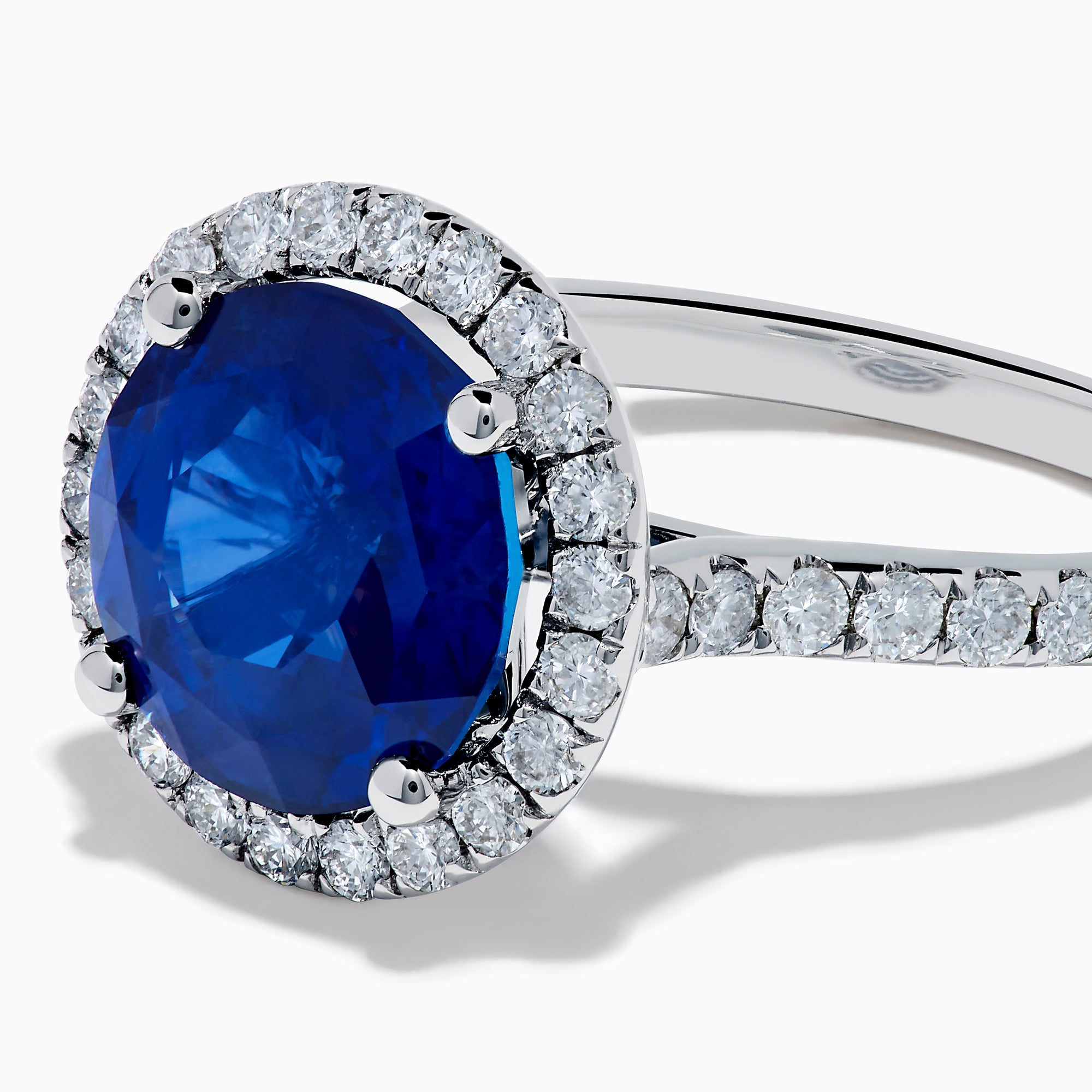 The Royale Blue 18K White Gold Blue Sapphire and Diamond Ring features a striking round blue sapphire center, encircled by a halo of sparkling diamonds, displayed against a white background.