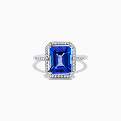 The Nahla Siri 14K White Gold Tanzanite and Diamond Ring features a large emerald-cut blue tanzanite, haloed by small white diamonds with additional diamonds set along the band.