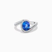 The 14K White Gold Tanzanite and Diamond Oval Swirl Ring features an oval tanzanite center stone accented by sparkling diamonds, all set on a modern, asymmetrical swirl band.