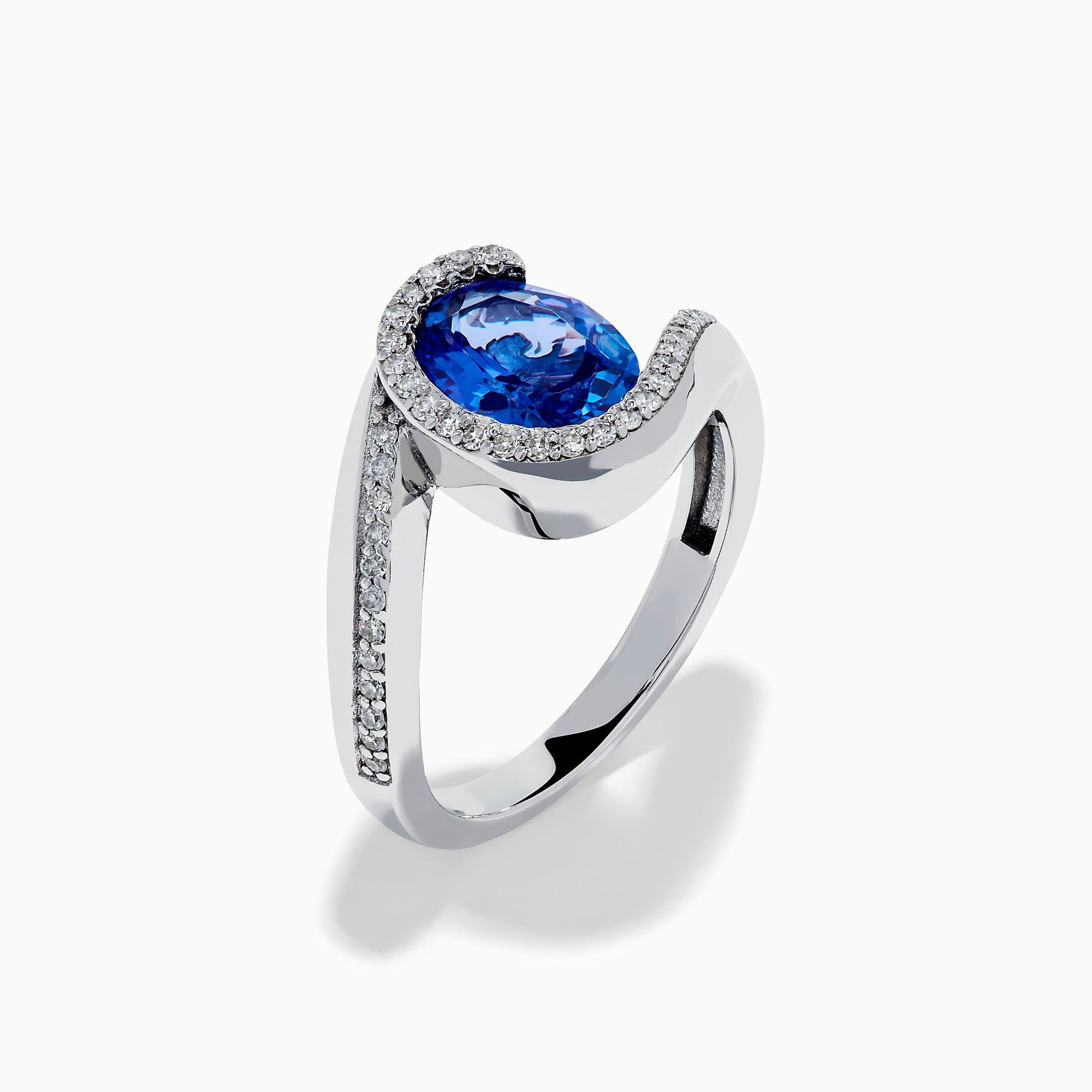 The 14K White Gold Tanzanite and Diamond Oval Swirl Ring features an oval tanzanite center stone accented by sparkling diamonds, all set on a modern, asymmetrical swirl band.