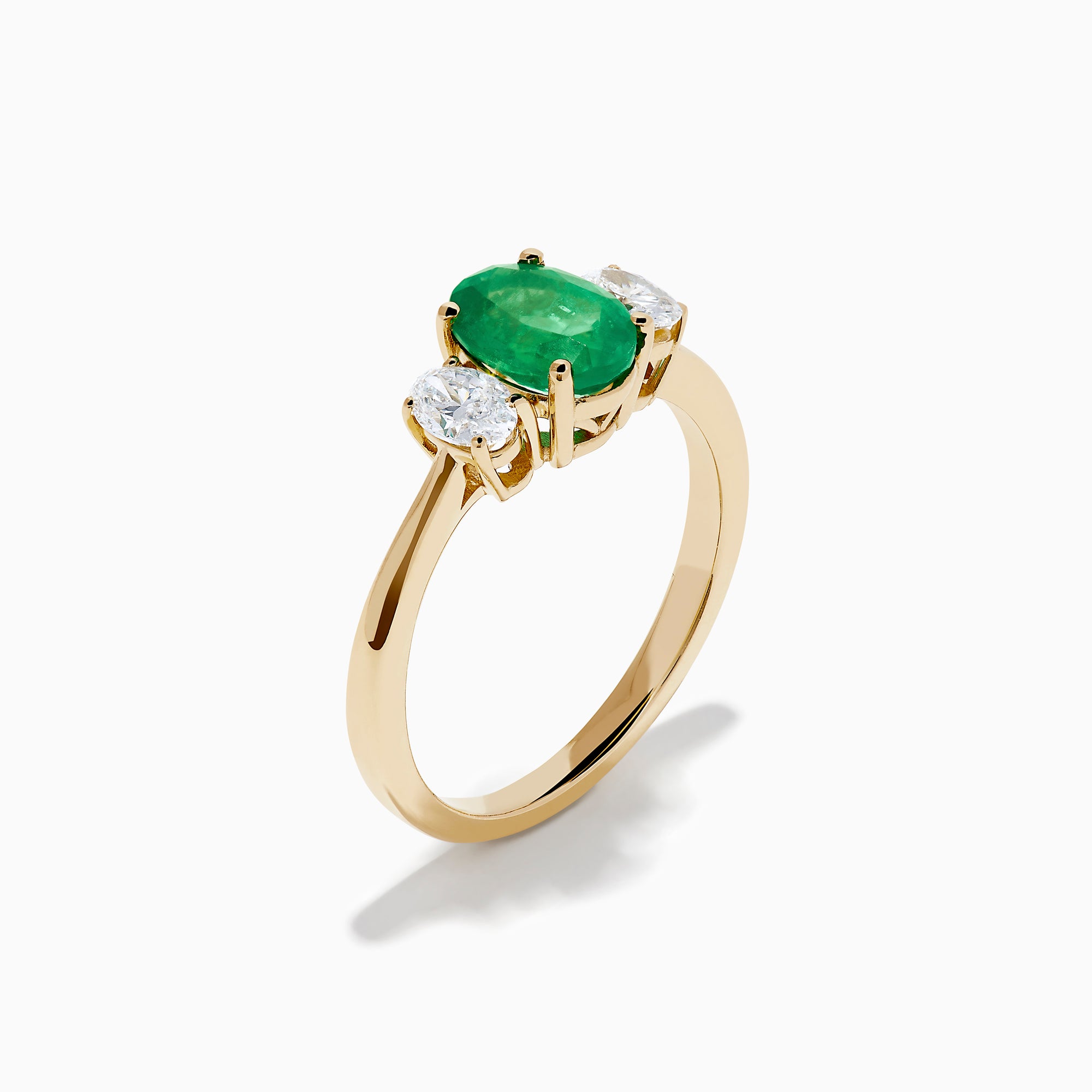 The 14K Yellow Gold Emerald and Diamond Three-Stone Ring showcases an oval green emerald center with two oval white diamonds on each side, all set in 14K yellow gold and displayed against a white background.