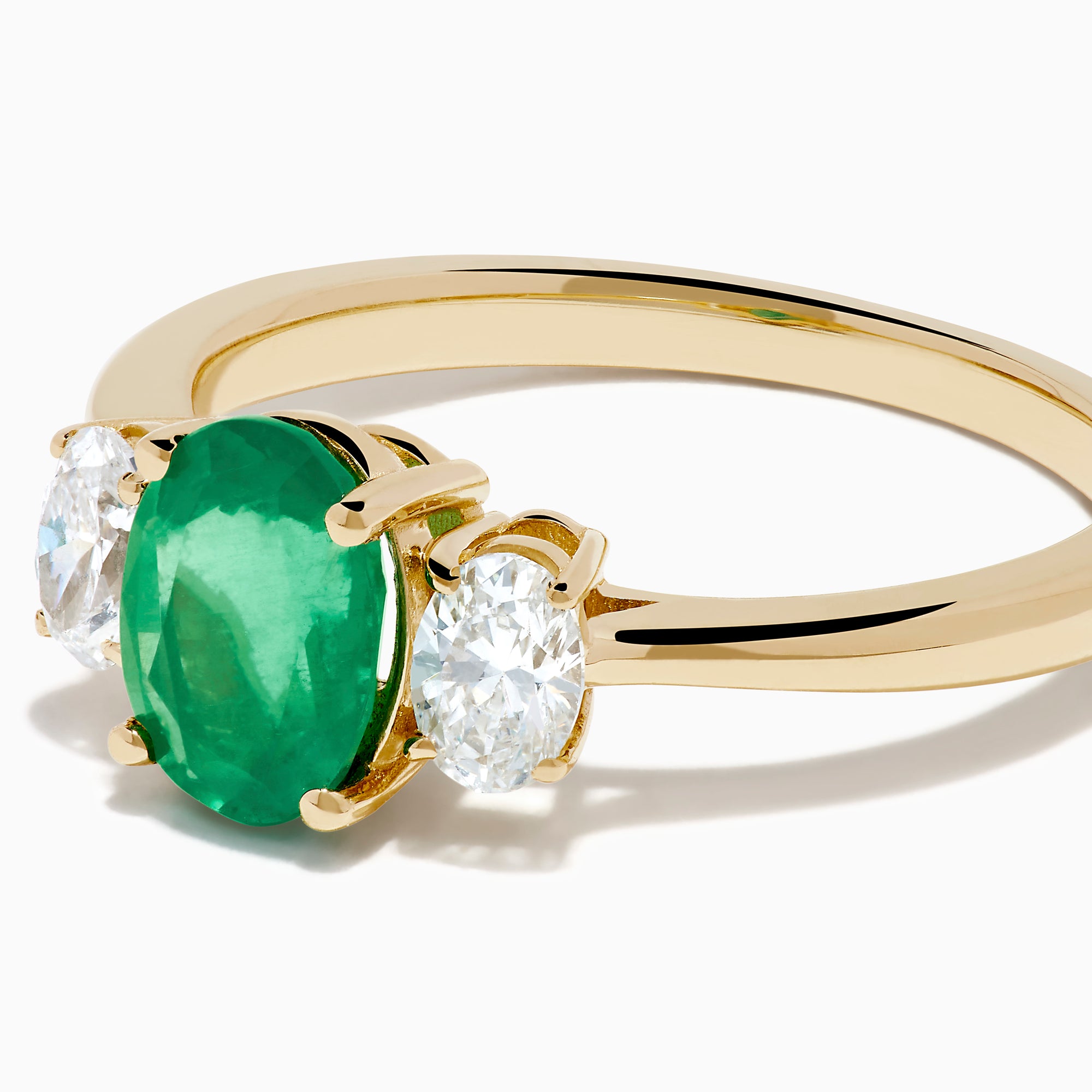 The 14K Yellow Gold Emerald and Diamond Three-Stone Ring showcases an oval green emerald center with two oval white diamonds on each side, all set in 14K yellow gold and displayed against a white background.