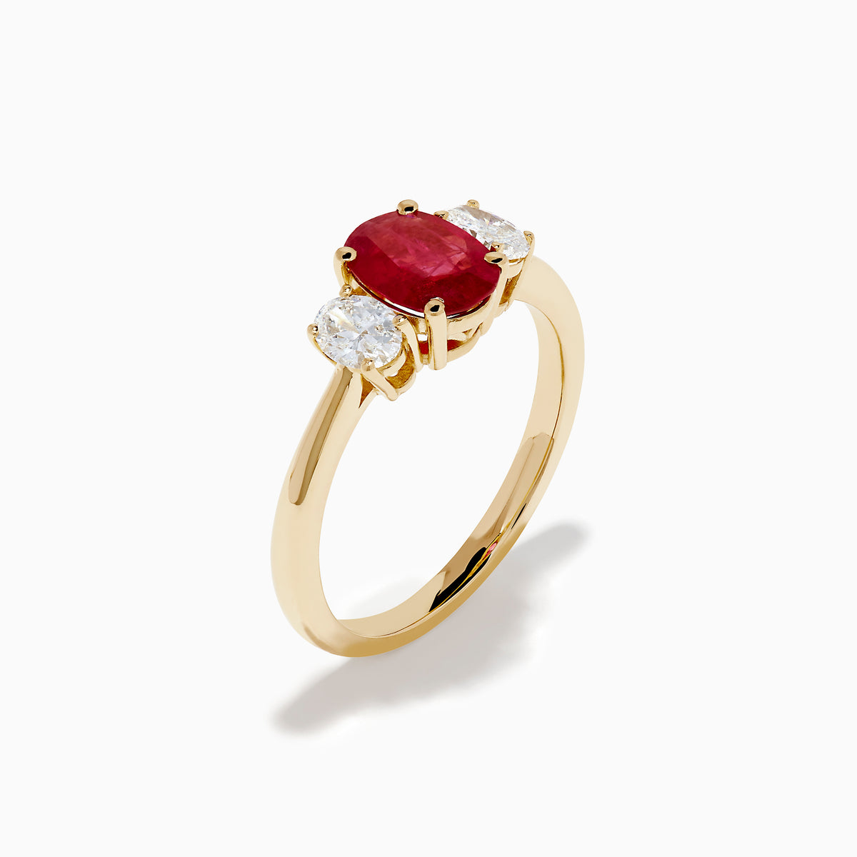 The 14K Yellow Gold Ruby and Diamond Three-Stone Ring showcases a striking oval ruby center with two oval white diamonds, set in yellow gold and displayed on a white background.