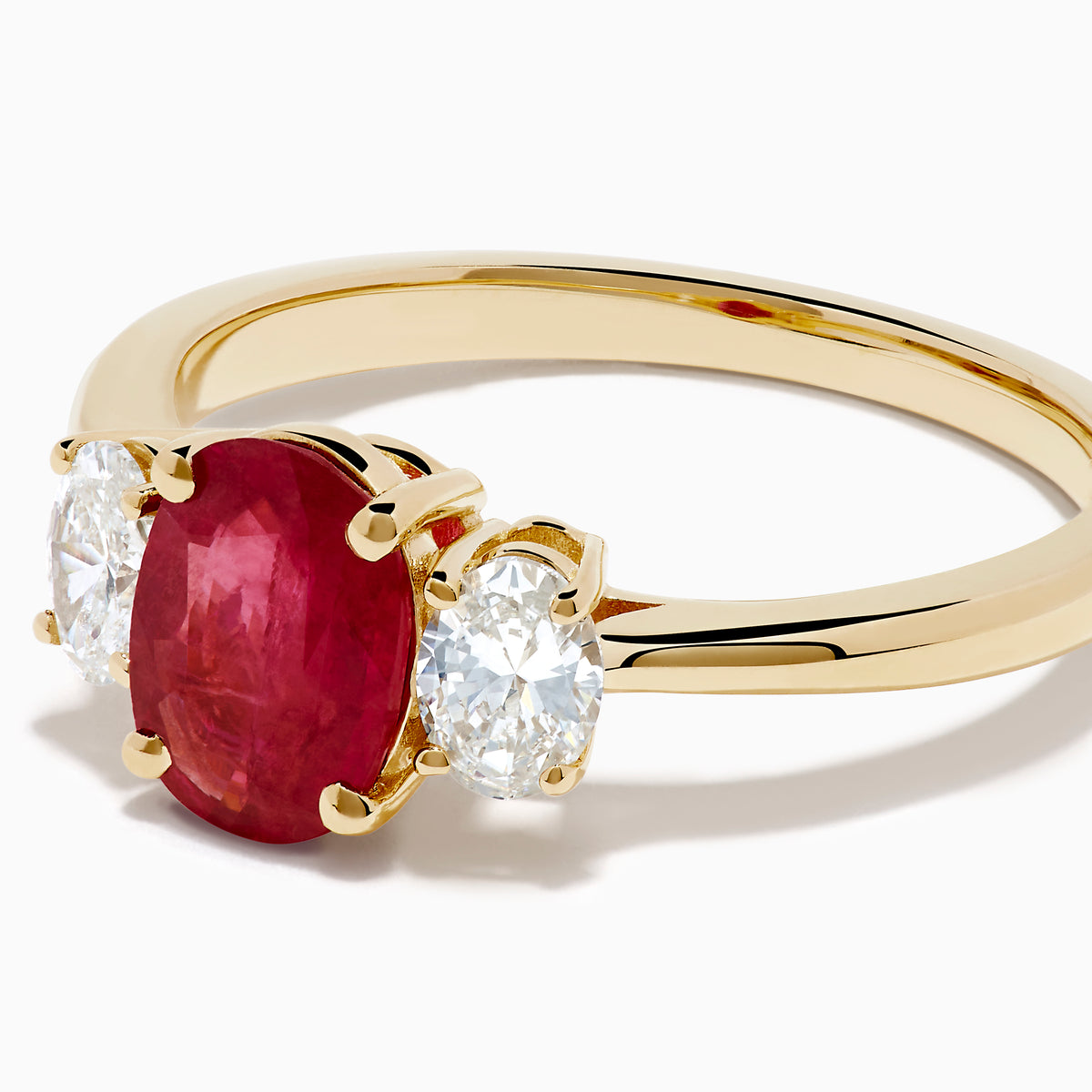 The 14K Yellow Gold Ruby and Diamond Three-Stone Ring showcases a striking oval ruby center with two oval white diamonds, set in yellow gold and displayed on a white background.