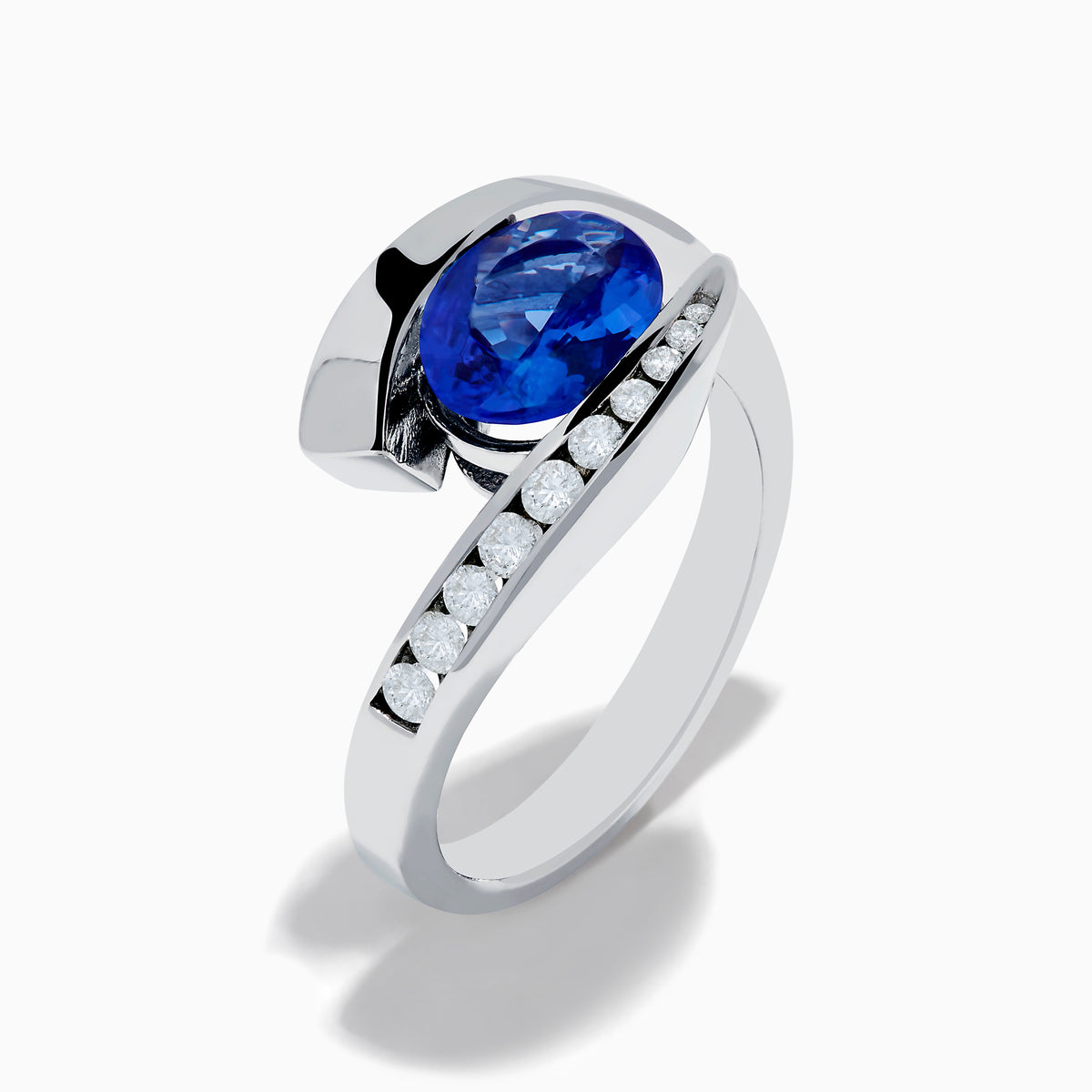 The Nahla 14K White Gold Floating Tanzanite and Diamond Ring showcases an oval blue tanzanite center with a curved band of round diamonds, all set in white gold and displayed on a white background.