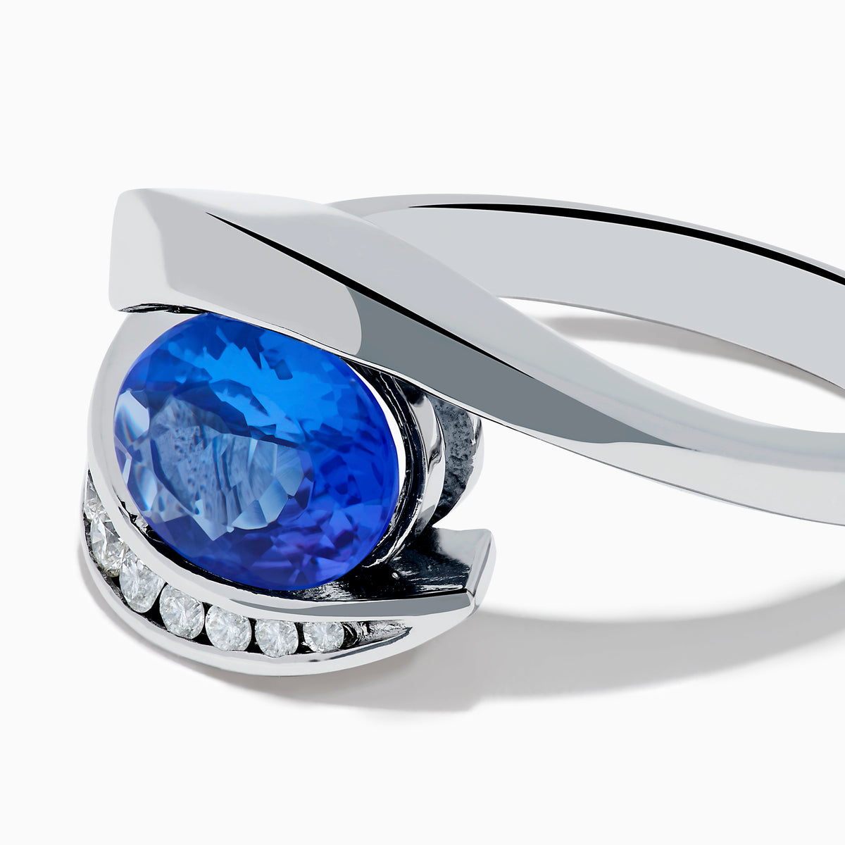The Nahla 14K White Gold Floating Tanzanite and Diamond Ring showcases an oval blue tanzanite center with a curved band of round diamonds, all set in white gold and displayed on a white background.