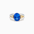 A striking 18K two tone gold ring featuring a large oval blue tanzanite center and two pear-shaped yellow diamonds on each side, displayed against a plain white background.