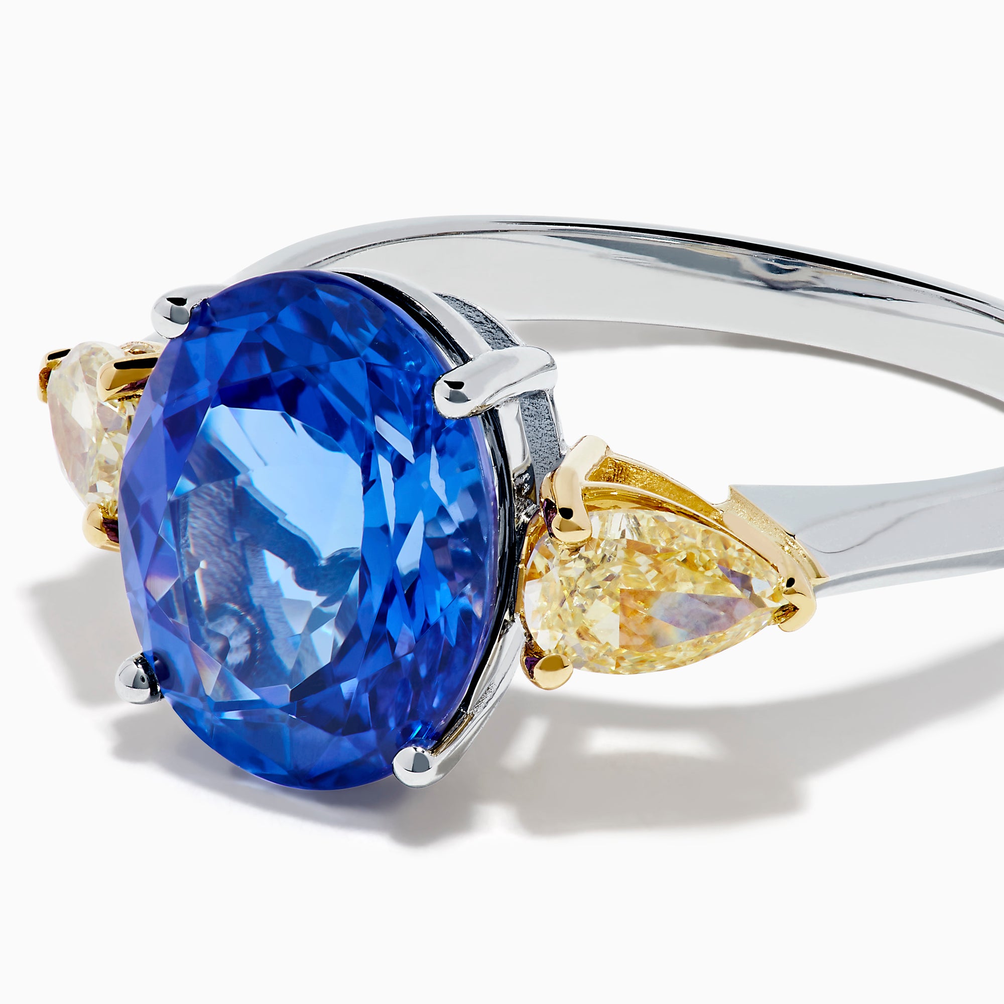 A striking 18K two tone gold ring featuring a large oval blue tanzanite center and two pear-shaped yellow diamonds on each side, displayed against a plain white background.