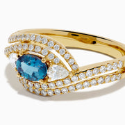 The 18K Yellow Gold Alexandrite and Diamond Swirl Halo Ring features a central oval blue alexandrite, two marquise-cut diamonds, and rows of small round diamonds in an eye-shaped swirl design.