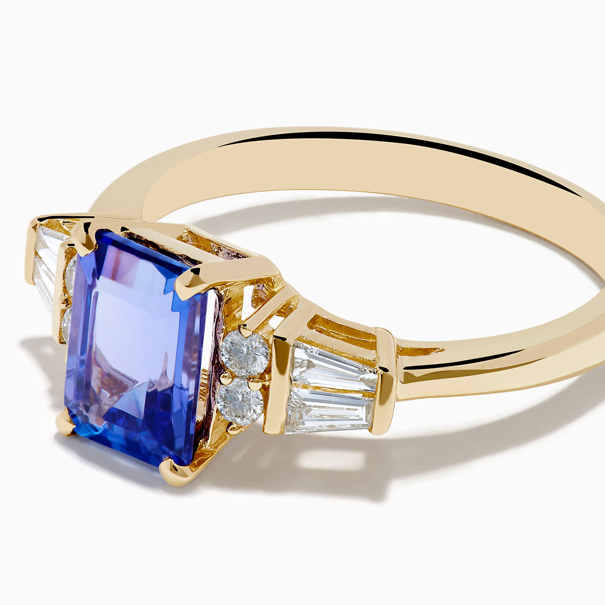 The Nahla 14K Yellow Gold Tanzanite and Diamond Ring showcases a rectangular blue tanzanite center, with two round diamonds and two baguette-cut white stones on each side, displayed on a white background.