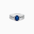 The 14K White Gold Blue Sapphire and Diamond Ring showcases an oval blue sapphire center, accented by two rows of round diamonds on each side, elegantly set against a white background.