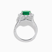 The 18K White Gold Emerald and Diamond Halo Ring features a large round green emerald center, surrounded by sparkling diamonds on the halo and band, beautifully displayed on a white background.