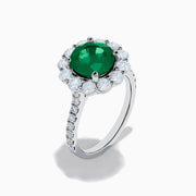 The 18K White Gold Emerald and Diamond Halo Ring features a large round green emerald center, surrounded by sparkling diamonds on the halo and band, beautifully displayed on a white background.