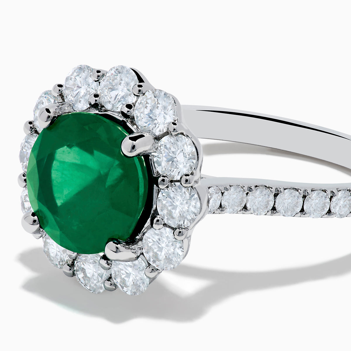 The 18K White Gold Emerald and Diamond Halo Ring features a large round green emerald center, surrounded by sparkling diamonds on the halo and band, beautifully displayed on a white background.