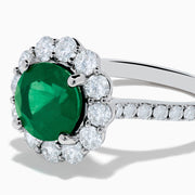 The 18K White Gold Emerald and Diamond Halo Ring features a large round green emerald center, surrounded by sparkling diamonds on the halo and band, beautifully displayed on a white background.