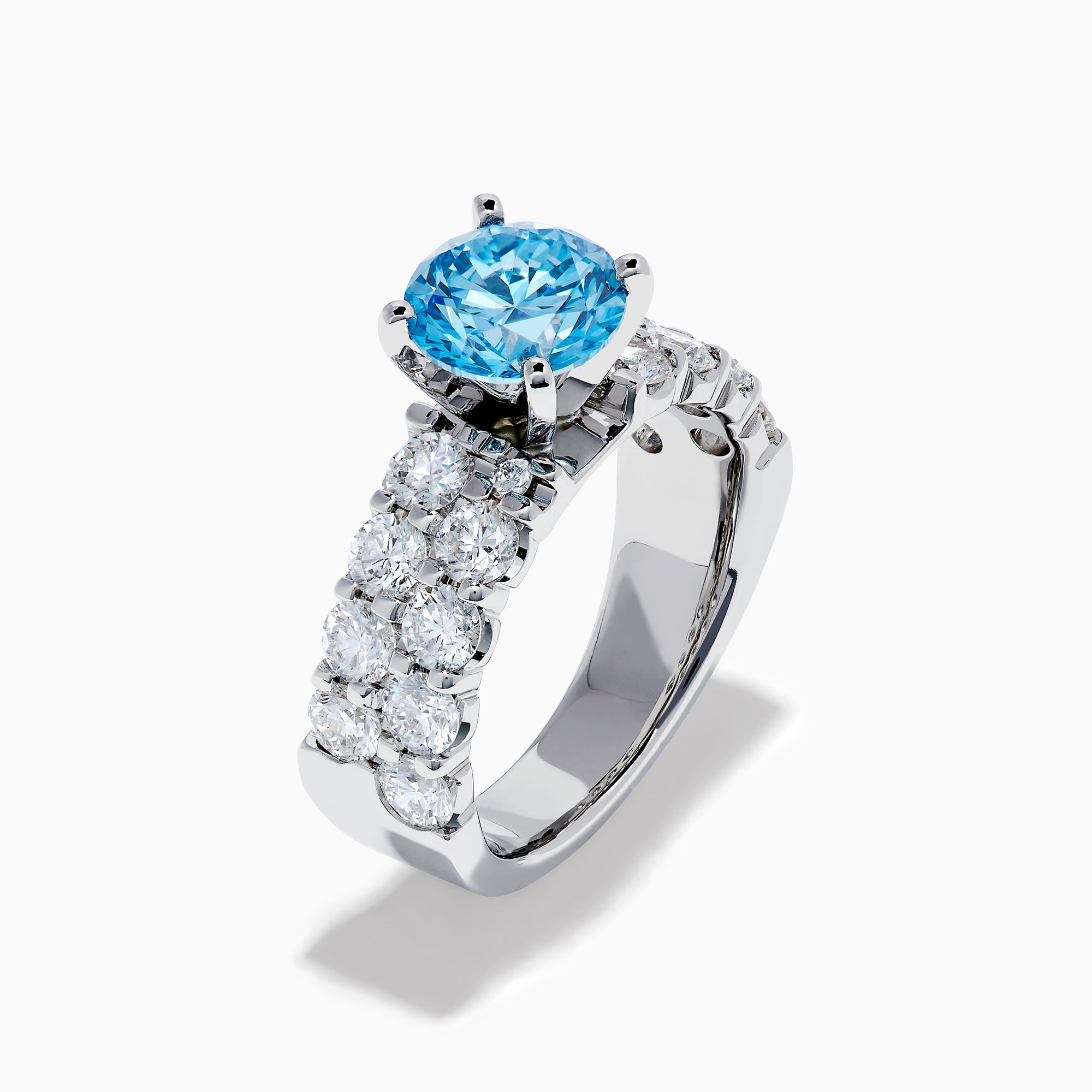 The Lab Grown Blue Diamond Round Brilliant Solitaire Ring in 14K White Gold features a striking round blue diamond center, accented by two rows of sparkling clear diamonds along the band, shown on a white background.