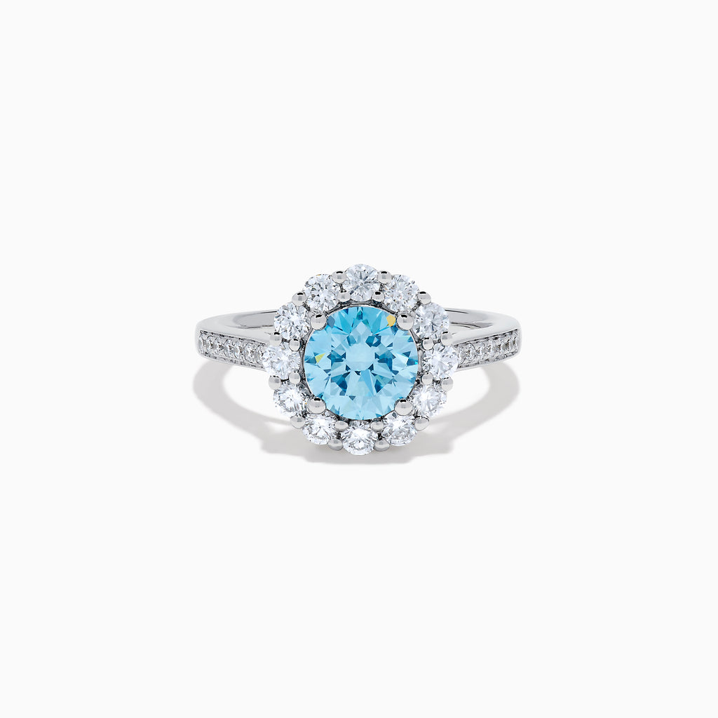The Lab Grown Blue and White Diamond Halo Ring in 14K White Gold showcases a striking round blue center stone, encircled by lab grown diamond accents and additional diamonds adorning the band.
