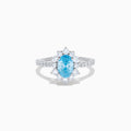 The Lab Grown Blue and White Diamond Halo Ring in 14K White Gold features an oval blue gemstone surrounded by lab grown diamonds, with more diamonds set along the band, shown on a white background.