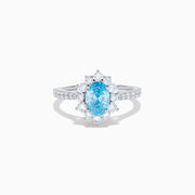 The Lab Grown Blue and White Diamond Halo Ring in 14K White Gold features an oval blue gemstone surrounded by lab grown diamonds, with more diamonds set along the band, shown on a white background.