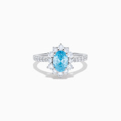 The Lab Grown Blue and White Diamond Halo Ring in 14K White Gold features an oval blue gemstone surrounded by lab grown diamonds, with more diamonds set along the band, shown on a white background.