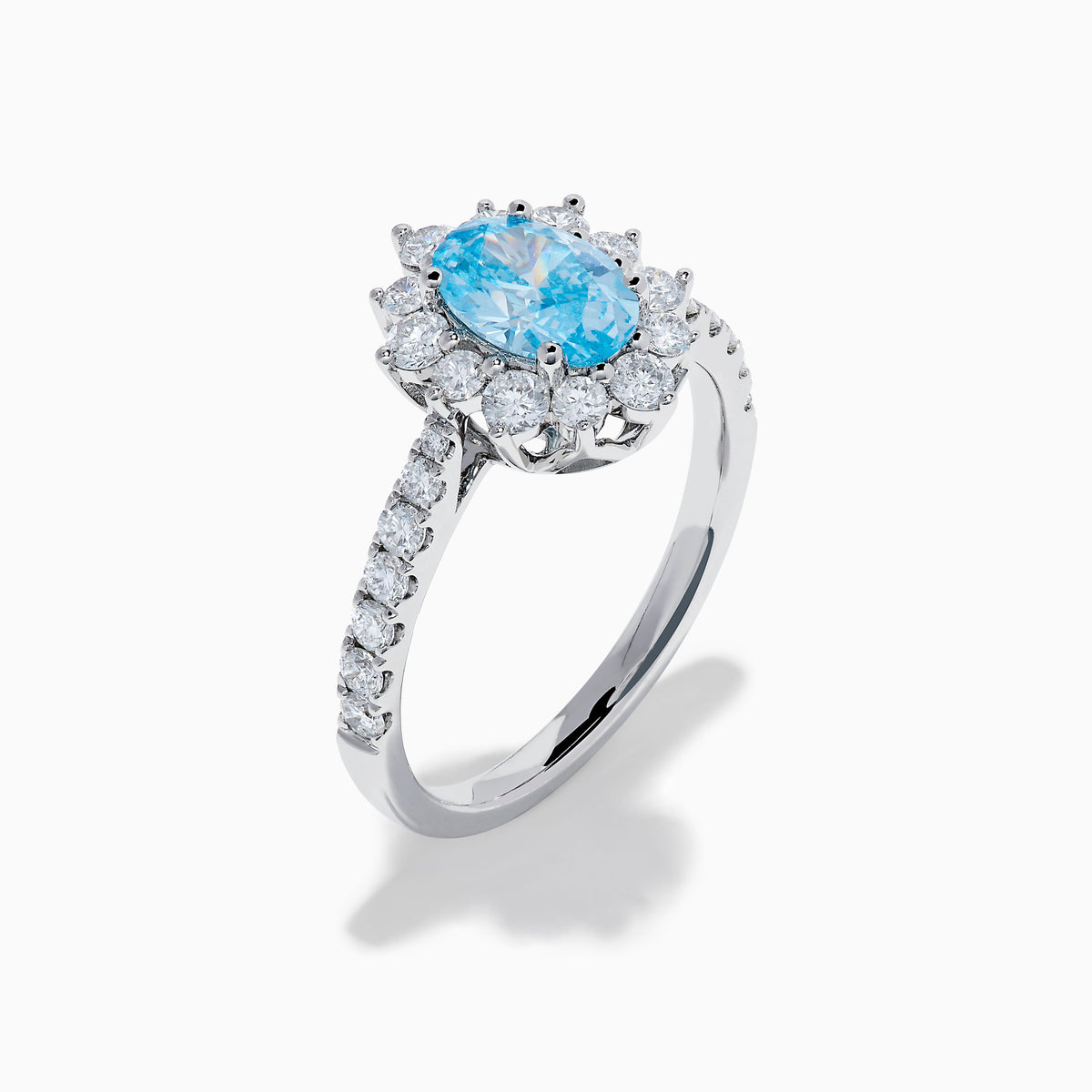 The Lab Grown Blue and White Diamond Halo Ring in 14K White Gold features an oval blue gemstone surrounded by lab grown diamonds, with more diamonds set along the band, shown on a white background.