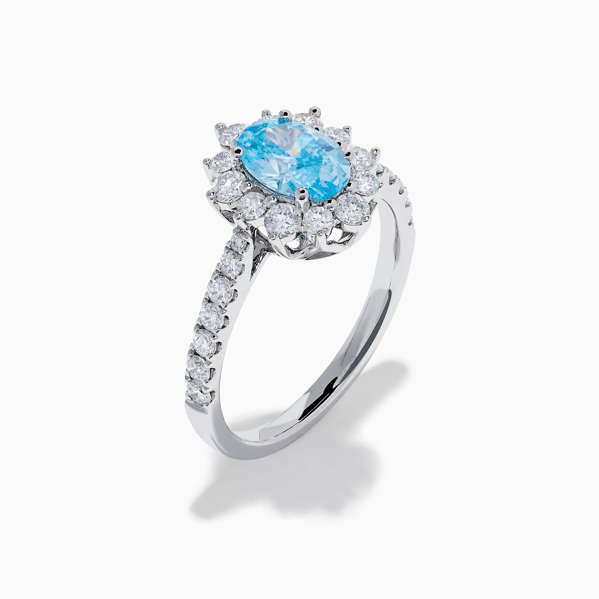 The Lab Grown Blue and White Diamond Halo Ring in 14K White Gold features an oval blue gemstone surrounded by lab grown diamonds, with more diamonds set along the band, shown on a white background.
