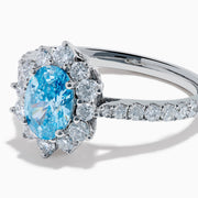 The Lab Grown Blue and White Diamond Halo Ring in 14K White Gold features an oval blue gemstone surrounded by lab grown diamonds, with more diamonds set along the band, shown on a white background.