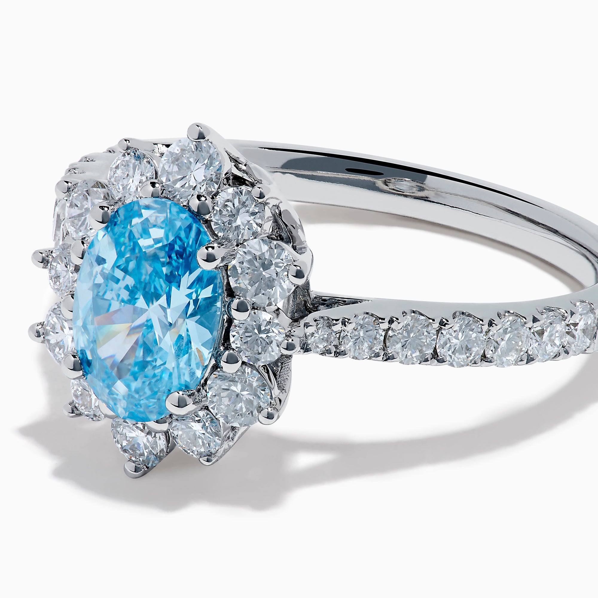 The Lab Grown Blue and White Diamond Halo Ring in 14K White Gold features an oval blue gemstone surrounded by lab grown diamonds, with more diamonds set along the band, shown on a white background.