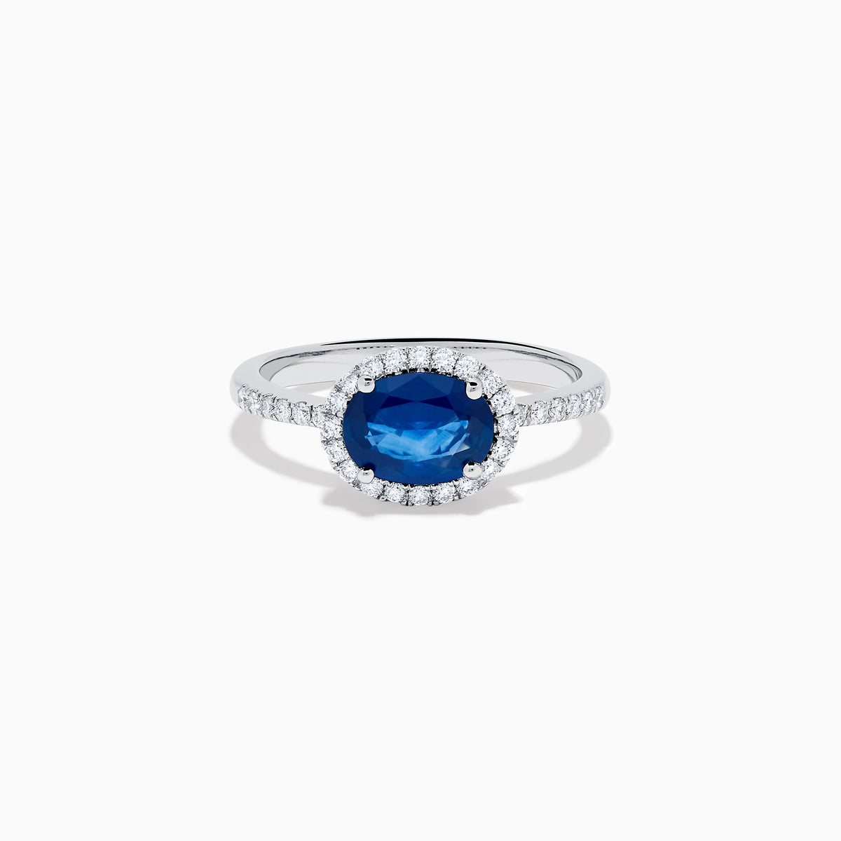 The 14K White Gold East West Oval Blue Sapphire and Diamond Halo Ring showcases an oval blue sapphire encircled by a diamond halo, with extra diamonds accenting the band for added brilliance.