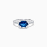 The 14K White Gold East West Oval Blue Sapphire and Diamond Halo Ring showcases an oval blue sapphire encircled by a diamond halo, with extra diamonds accenting the band for added brilliance.