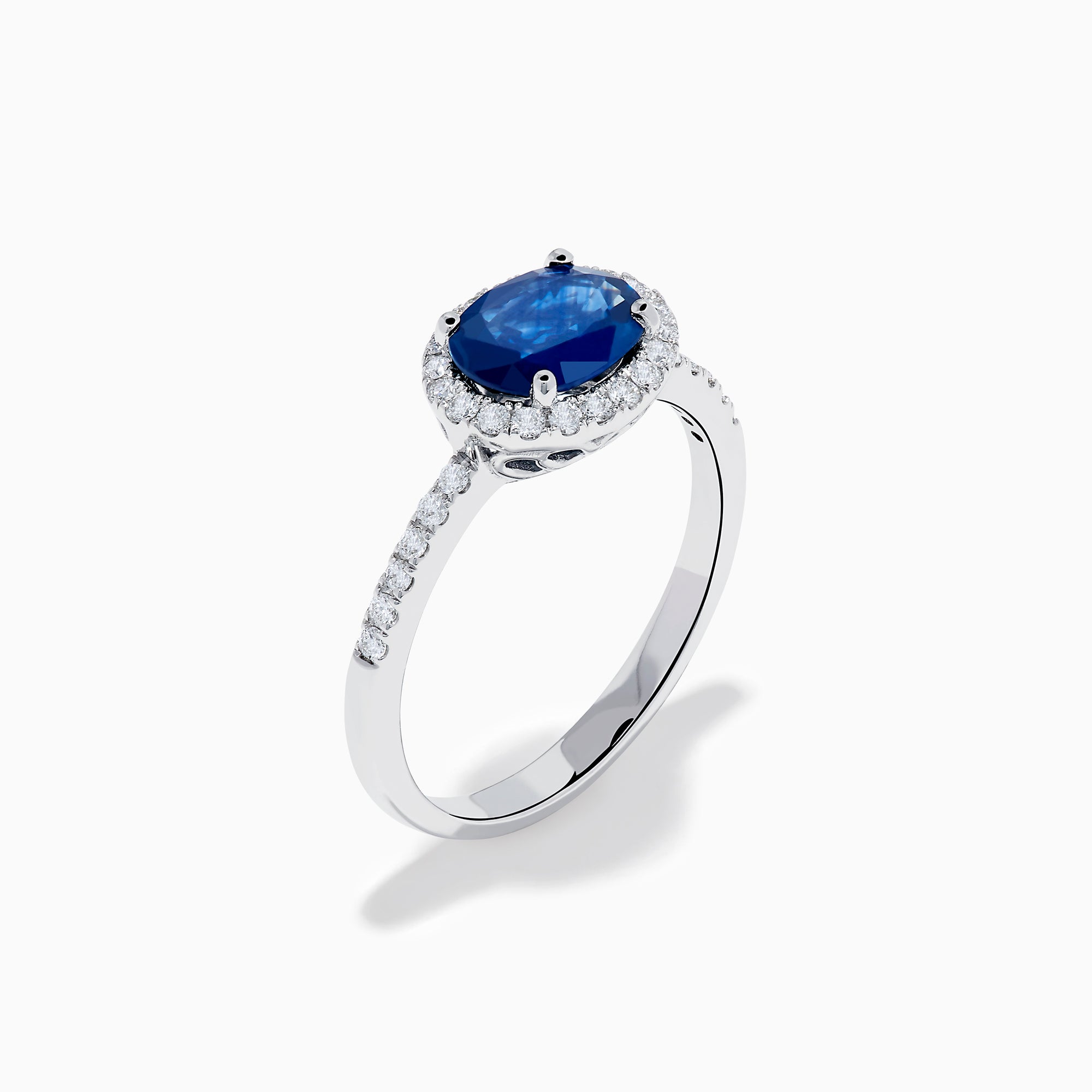 The 14K White Gold East West Oval Blue Sapphire and Diamond Halo Ring showcases an oval blue sapphire encircled by a diamond halo, with extra diamonds accenting the band for added brilliance.