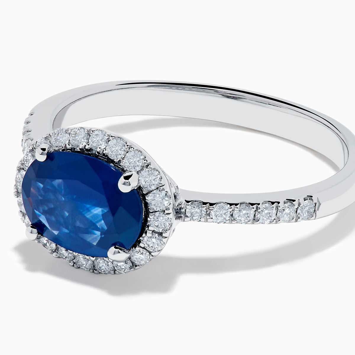 The 14K White Gold East West Oval Blue Sapphire and Diamond Halo Ring showcases an oval blue sapphire encircled by a diamond halo, with extra diamonds accenting the band for added brilliance.