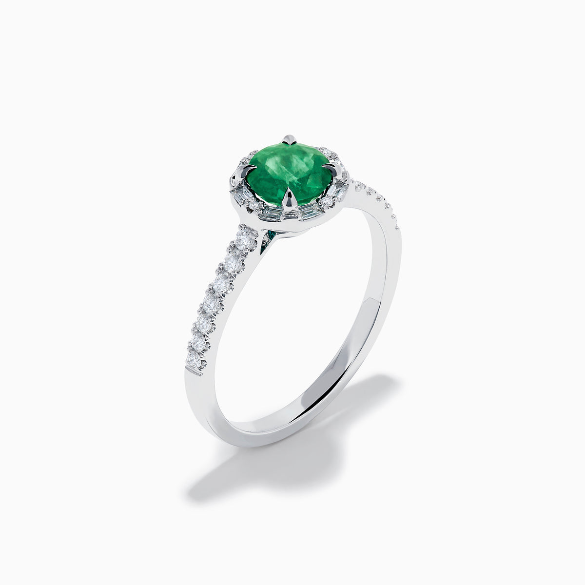 Introducing the 14K White Gold Emerald and Diamond Baguette Halo Ring, showcasing a round green emerald center stone, diamond baguette halo, and a band embellished with shimmering clear stones for added elegance.