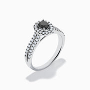 The 14K White Gold White and Black Diamond Halo Ring features an oval black center stone, a halo of white diamonds, and additional diamonds set along its split shank band.