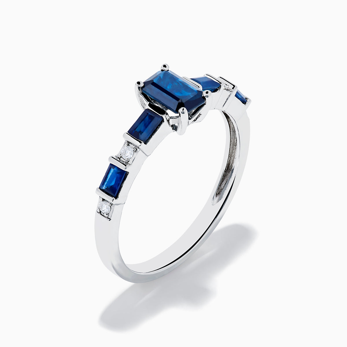 The Royal Bleu 14K White Gold Blue Sapphire and Diamond Ring features a rectangular blue sapphire center, flanked by square blue stones and diamonds on a sleek band against a plain white background.
