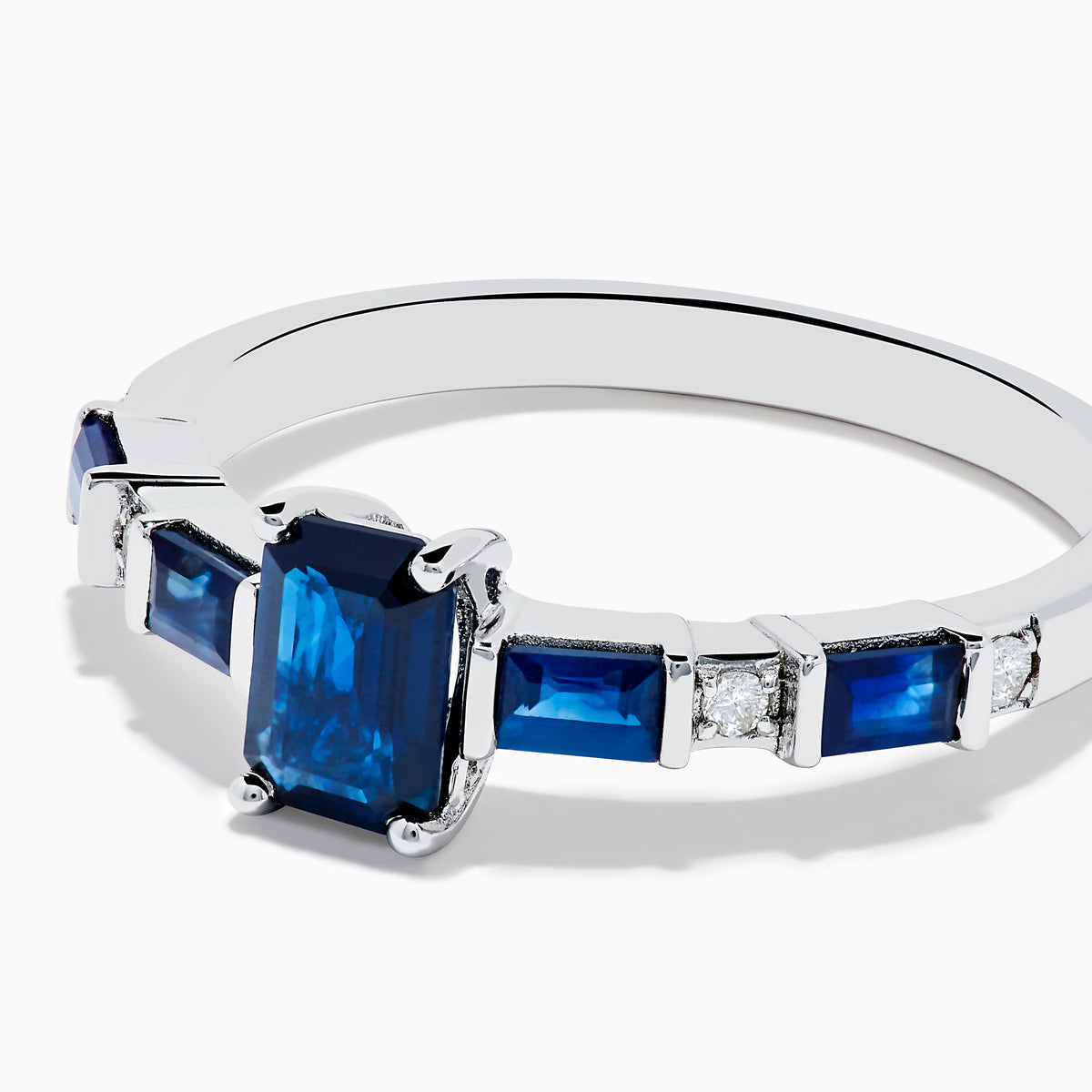 The Royal Bleu 14K White Gold Blue Sapphire and Diamond Ring features a rectangular blue sapphire center, flanked by square blue stones and diamonds on a sleek band against a plain white background.