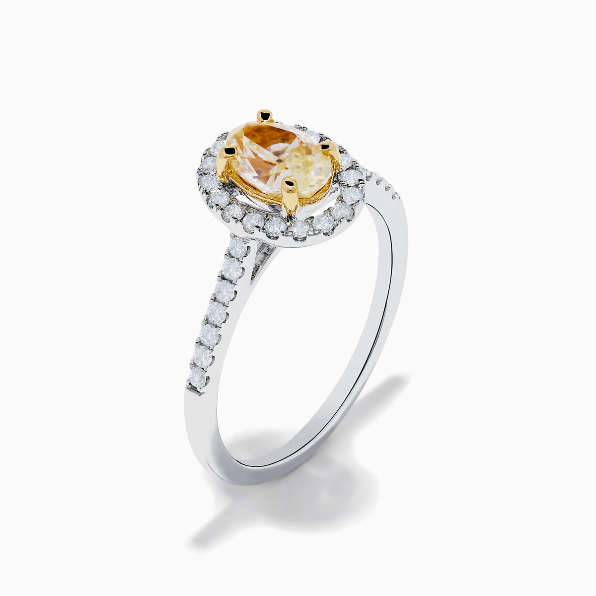 The Canare 18K Two Tone White and Yellow Diamond Ring features an oval yellow diamond center, surrounded by a halo of round white diamonds with extra diamonds adorning the band.