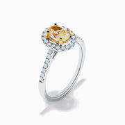 The Canare 18K Two Tone White and Yellow Diamond Ring features an oval yellow diamond center, surrounded by a halo of round white diamonds with extra diamonds adorning the band.