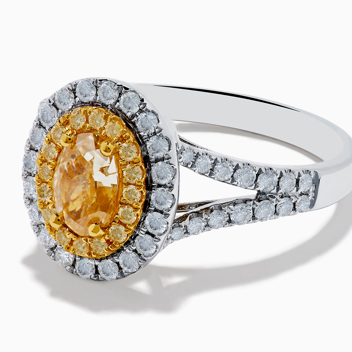The Canare 18K Two Tone Gold White and Yellow Oval Diamond Split Shank Ring showcases an oval yellow diamond center with a double halo of white diamonds and a diamond-accented split shank, displayed on a plain white background.